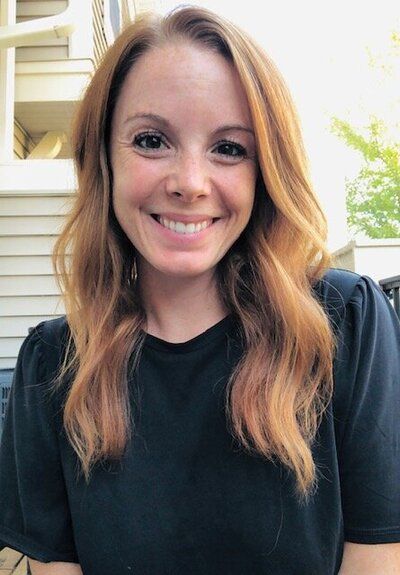 Woman with auburn hair smiles, wearing a black shirt, outside.