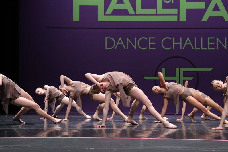 Dancers in brown costumes perform a dance, bending backward on a stage with a purple backdrop.