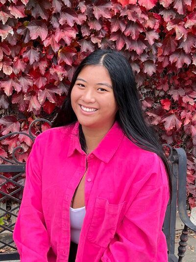Woman with long dark hair, wearing a pink shirt, smiles in front of red fall foliage.