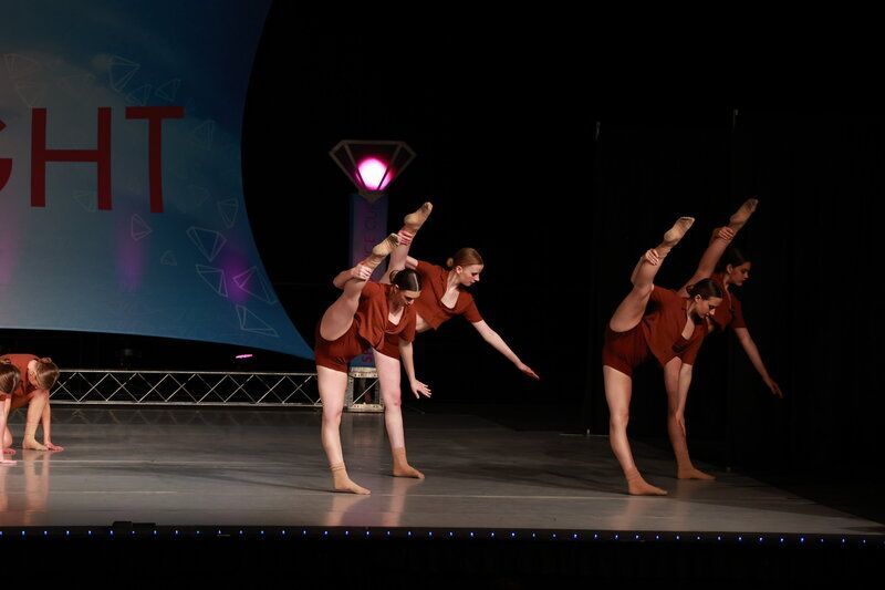 Dancers in brown costumes perform a synchronized pose on a stage, legs raised high, arms extended.