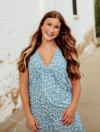 Woman in blue floral top and skirt, smiling, standing outside a white brick building.