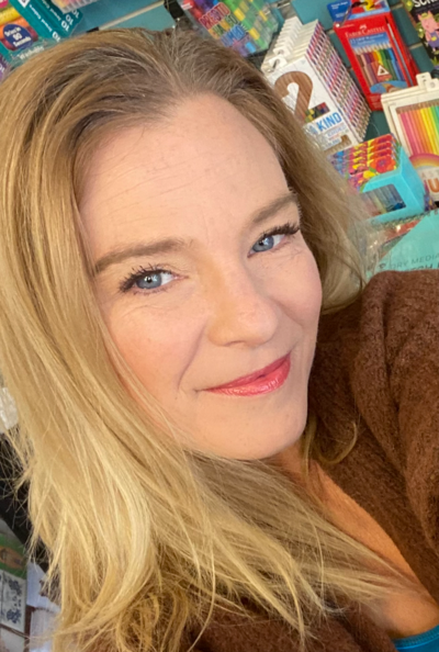 Woman with blonde hair smiles at the camera, wearing a brown sweater in a colorful setting.