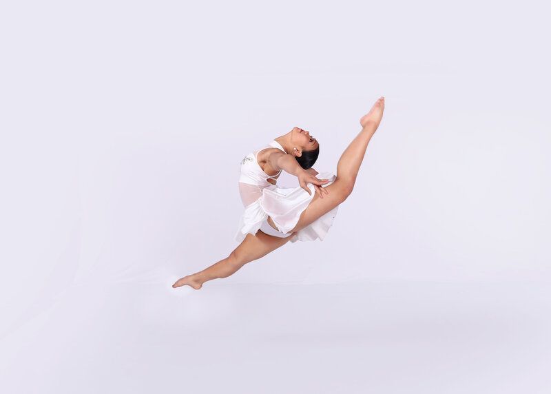 Dancer leaps high, in a split, arms overhead. She wears a white dress against a white background.