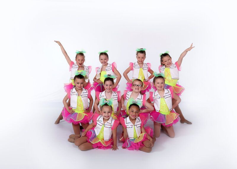 Dance group in bright costumes, posing in front of white background, arms raised, smiles.