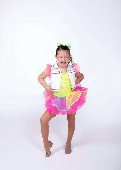 Young dancer in colorful tutu, posing with one leg raised and a smile, studio setting.