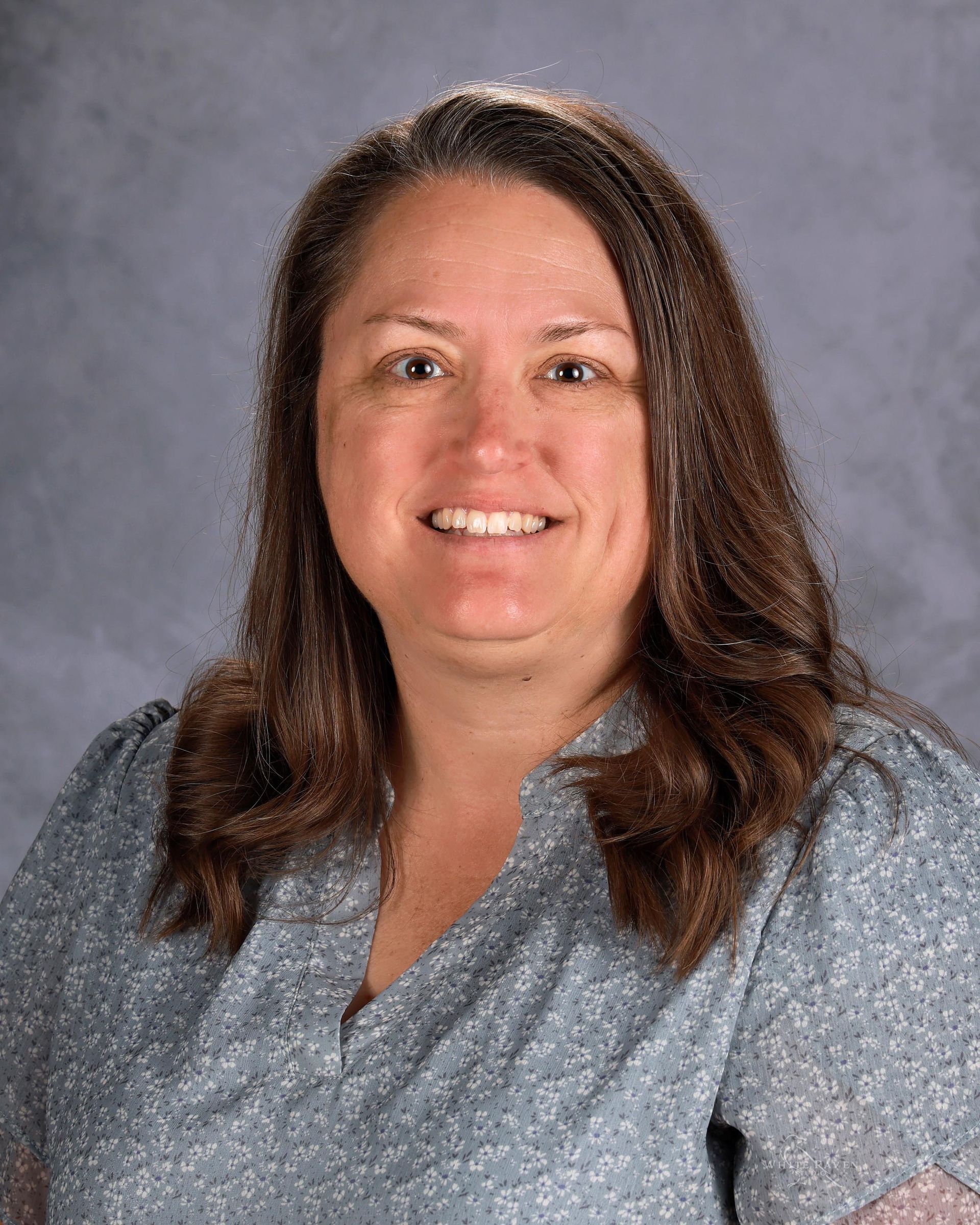 A woman wearing a blue shirt and a necklace is smiling for the camera.