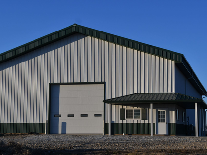 Large metal building with green trim, garage door, and small attached office under a blue sky.