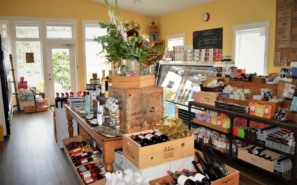 Interior of a cozy market with wooden display tables featuring wine bottles, gift items, and a bouquet, plus shelf goods.