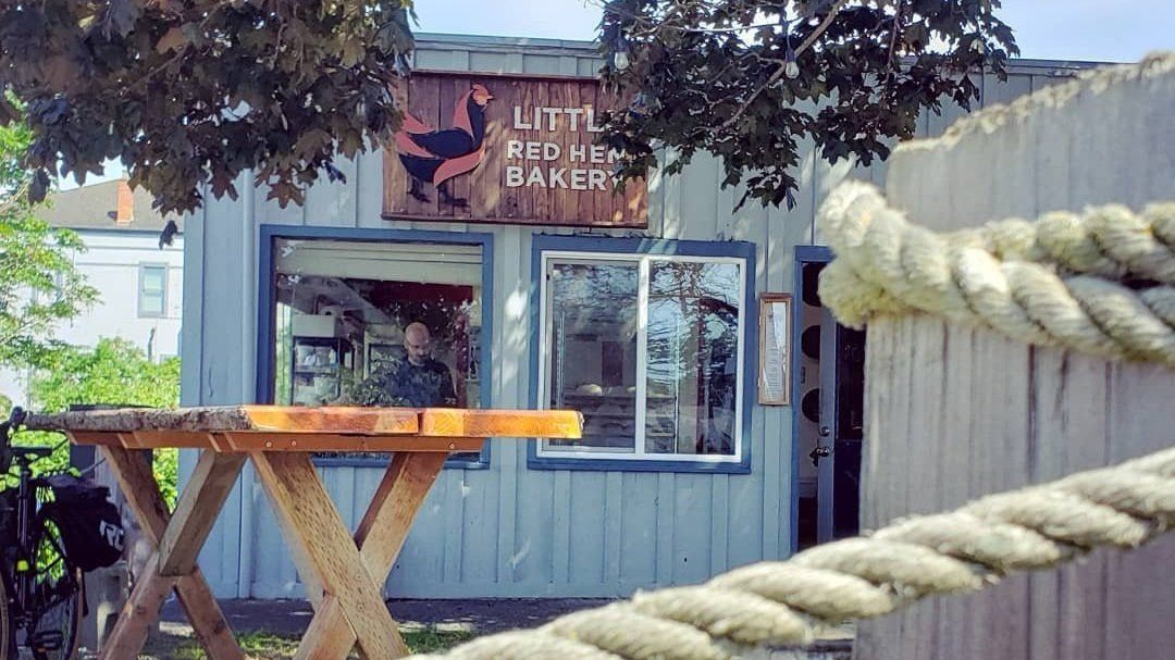 Little Red Hen Bakery storefront with a wooden table in front and a rope railing along the right side.