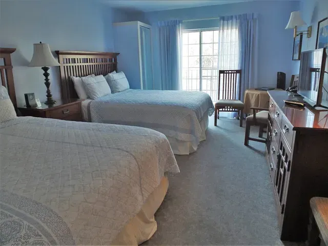 A bedroom with two beds, blue walls, a wooden dresser, a desk chair, and large windows with sheer curtains.