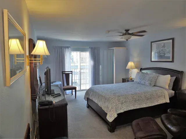 A bedroom featuring a queen bed with patterned bedding, dark furniture, a ceiling fan, and a sliding glass door to a balcony.