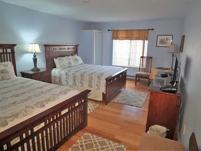 A bedroom with two wooden beds, a desk, a window with tan curtains, and blue walls, with patterned rugs on hardwood floors.