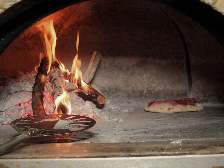 A wood-fired pizza oven with a burning fire on the left and a flat, raw dough disc cooking on the stone hearth.