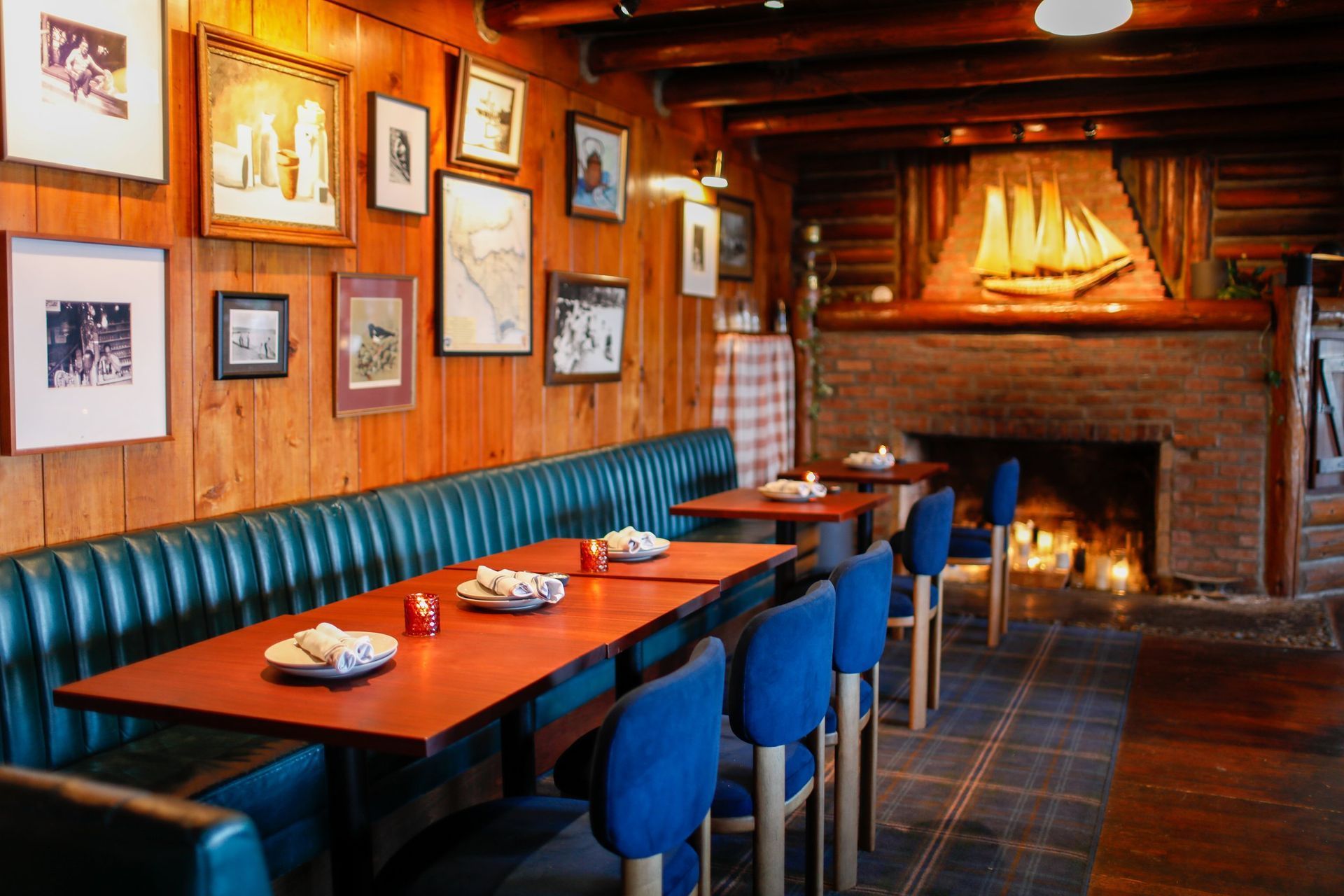 A rustic dining area with wooden walls, a fireplace featuring a ship art piece, and tables with blue chairs and booths.
