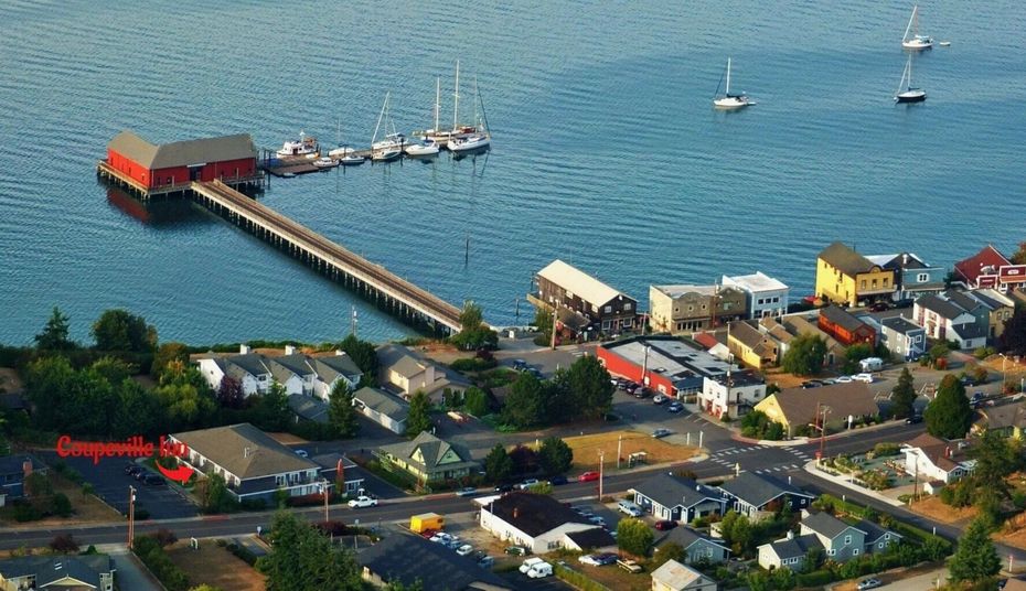 An aerial view of a coastal town with a long pier leading to a red building over the blue water, with moored sailboats.