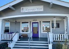 Lavender Wind shop with gray siding, a purple door, steps, and a railing under a covered porch.