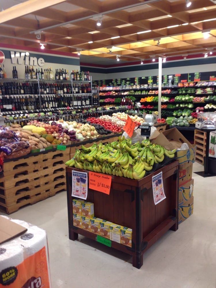 A grocery store produce section with a wooden display of green bananas in the foreground and shelves of wine and produce.