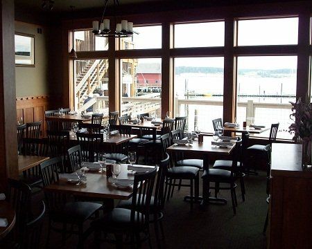 A dining area in a restaurant overlooking a pier and water through large windows, featuring wooden tables and chairs.