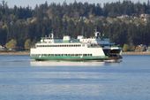 A white and green Washington State ferry cruising on calm water in front of a treelined shore.