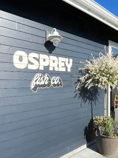 The exterior of Osprey Fish Co. featuring white text on a dark blue shingled wall with an outdoor light and a potted tree.