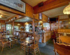 A rustic restaurant interior featuring wooden walls, tables with matching chairs, and a bar area with pendant lights.