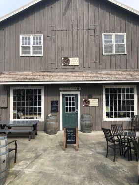 A wooden barn-style building with a green front door, patio seating, and a sidewalk sign in front.