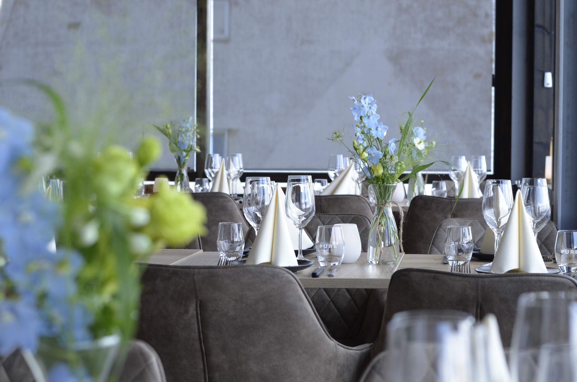 En restaurantinteriør med dækkede borde til spisning med blomsterarrangementer, glasvarer og stole i neutrale farver.