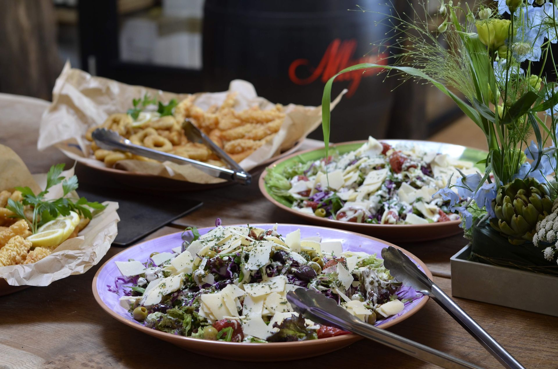 To salatskåle med ostespåner og en tallerken med friturestegt mad på et træbord med et blomsterarrangement i nærheden.