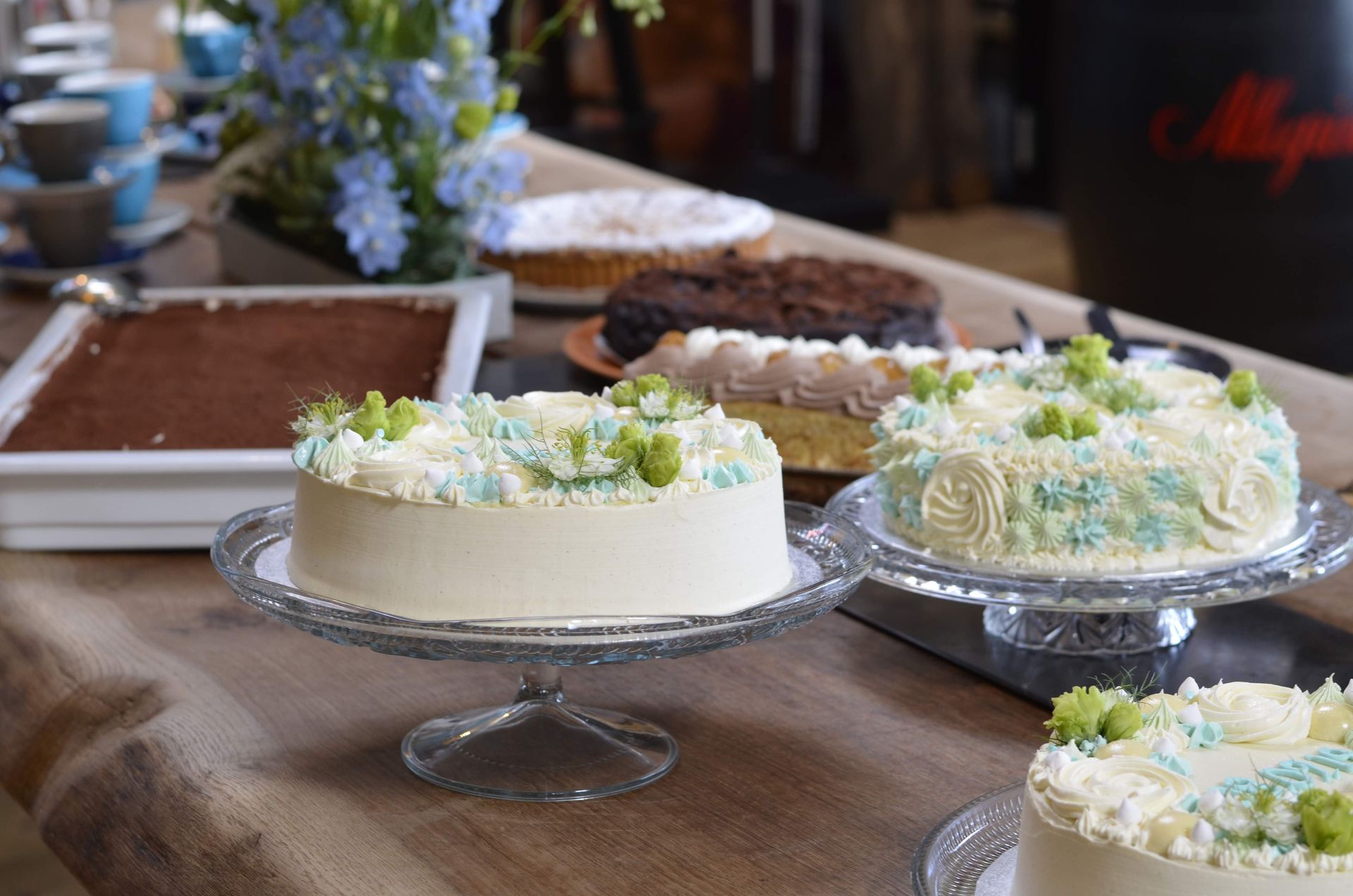 Adskillige kager, herunder dekorerede runde kager på fade og en rektangulær tiramisu, er arrangeret på et langt træbord.