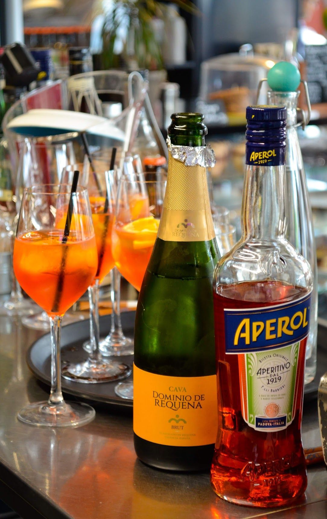 En flaske Aperol, en flaske mousserende vin og adskillige orange Aperol Spritz-cocktails på en bardisk.