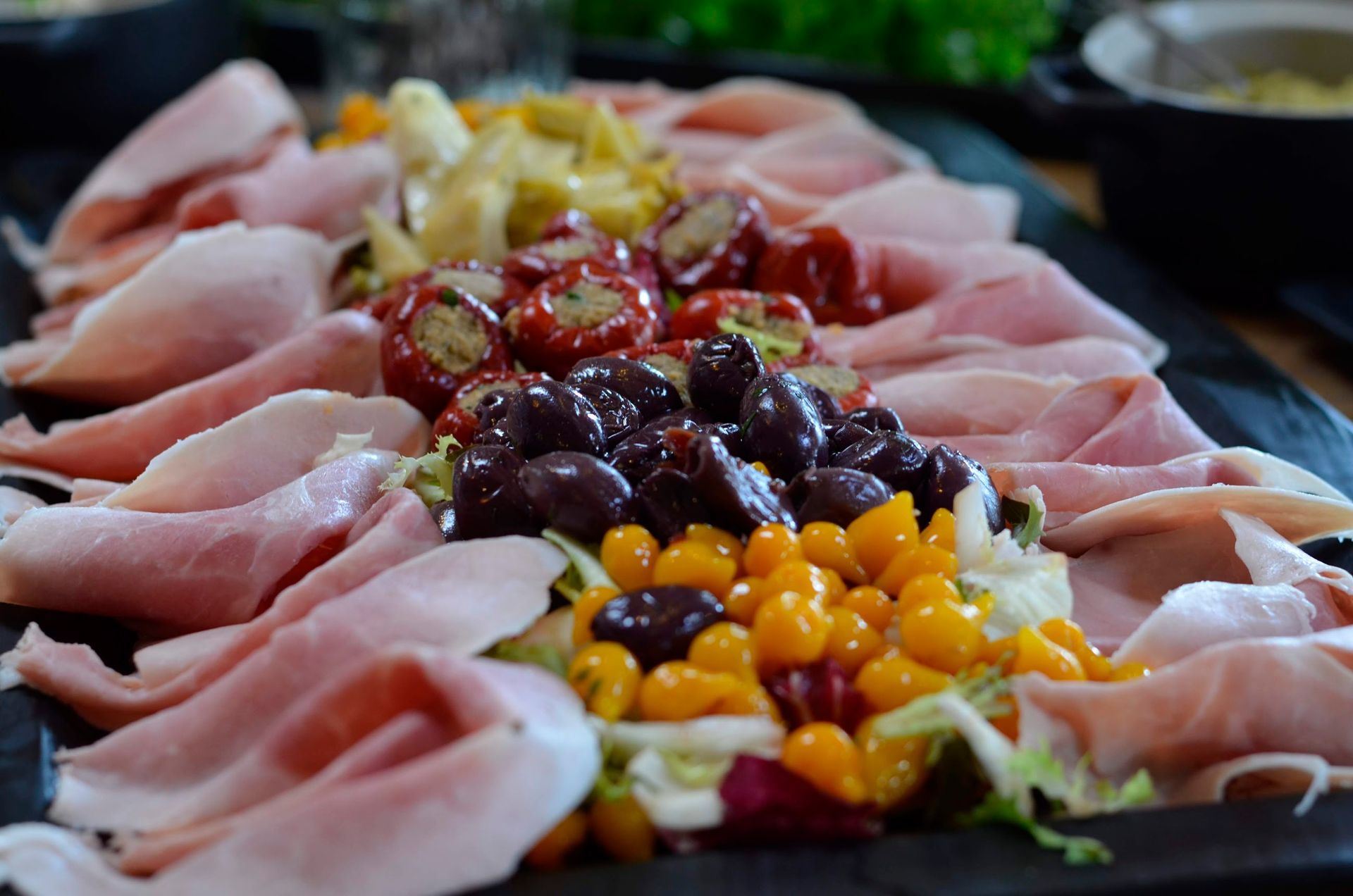 Et serveringsfad med foldede skinkeskiver omkranset af fyldte røde peberfrugter, sorte oliven og gule tomater.