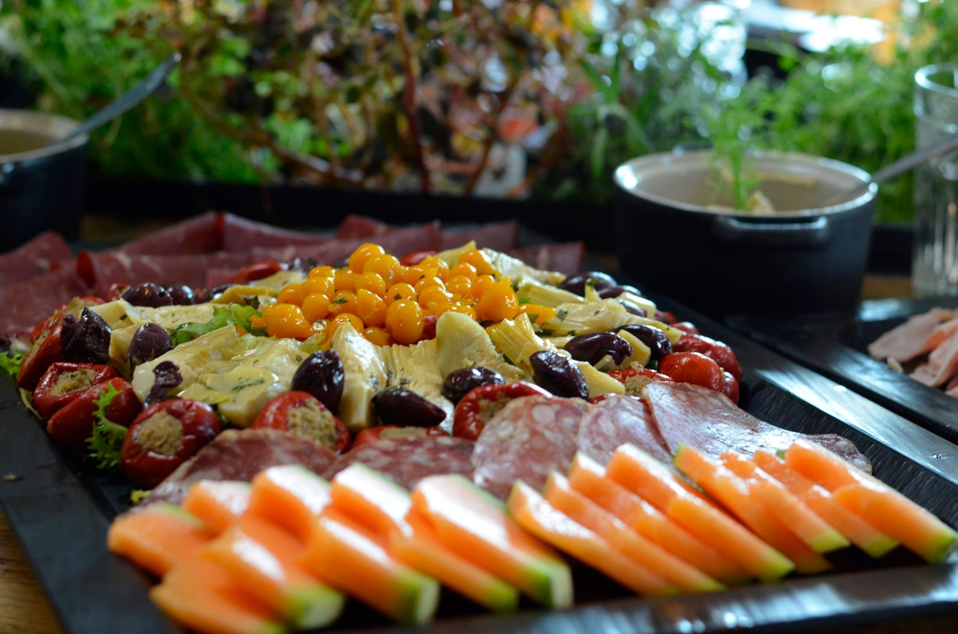 Et serveringsfad med charcuteri, melonskiver, oliven, artiskokker, fyldte peberfrugter og små gule tomater.