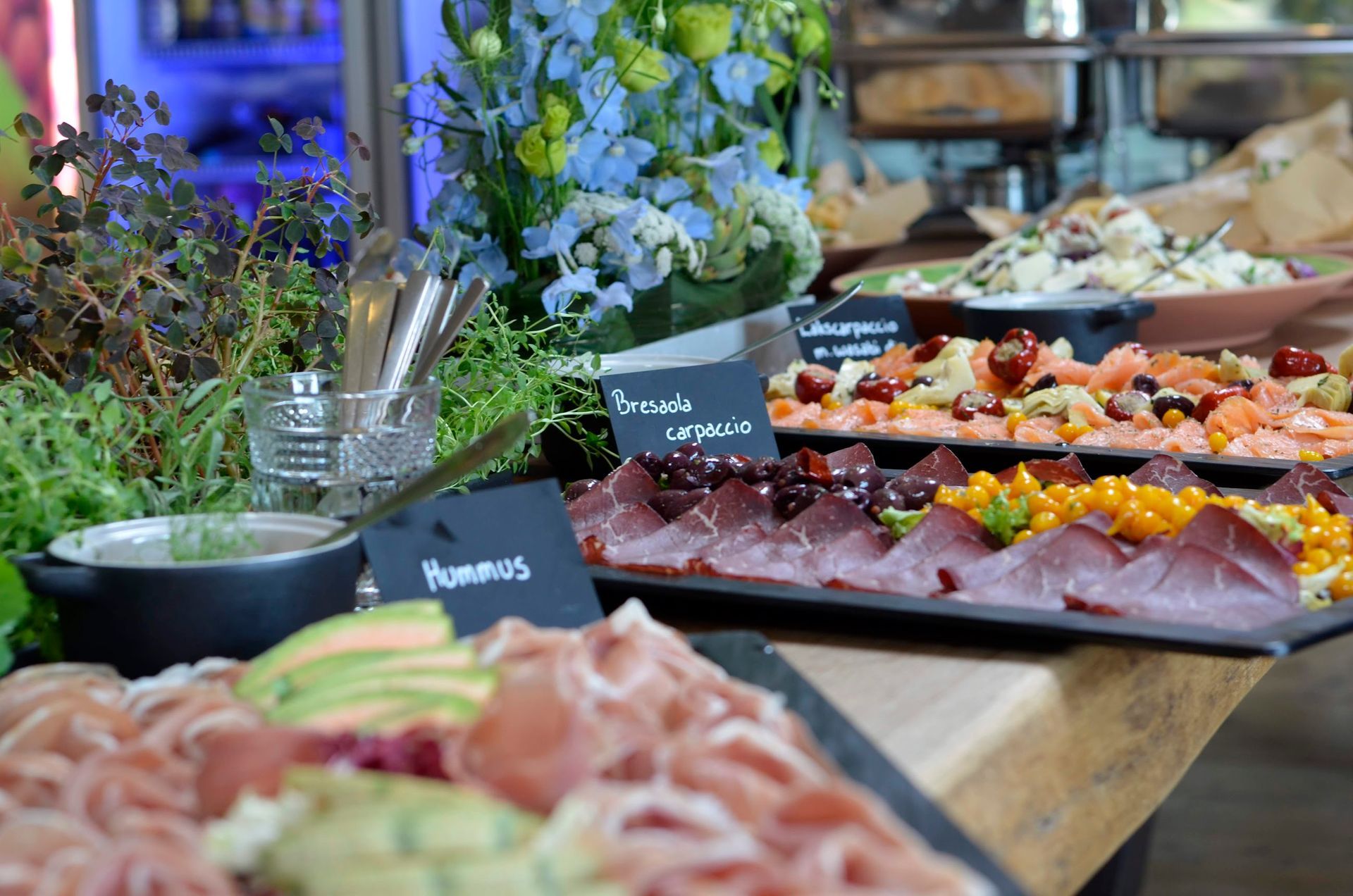 En buffet med forskellige charcuterier, friske grøntsager, hummus og salater, dekoreret med farverige blå blomster.