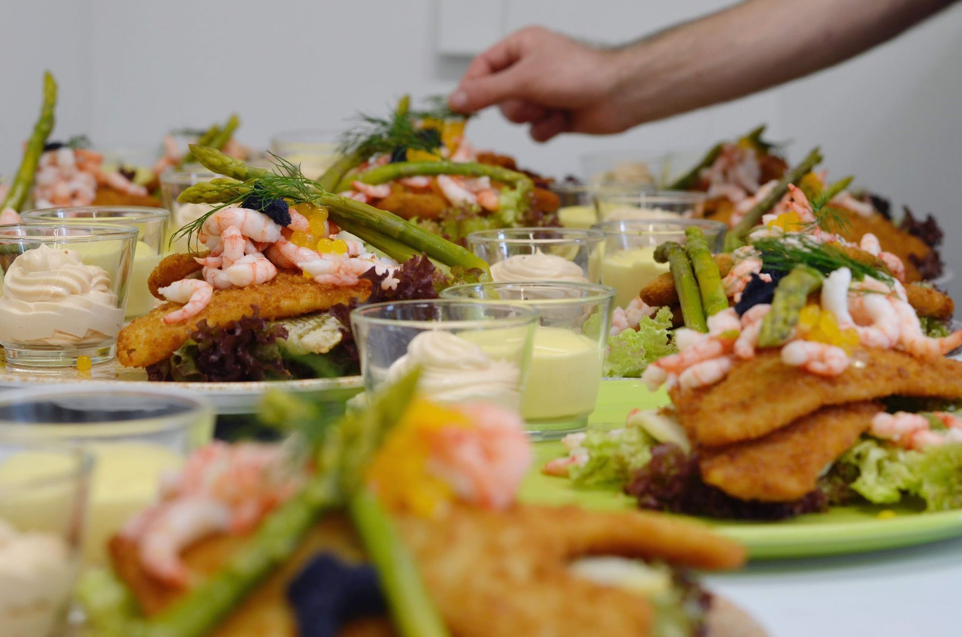 Tallerkener med paneret fisk toppet med rejer og asparges, med en hånd der pynter på den ene portion.