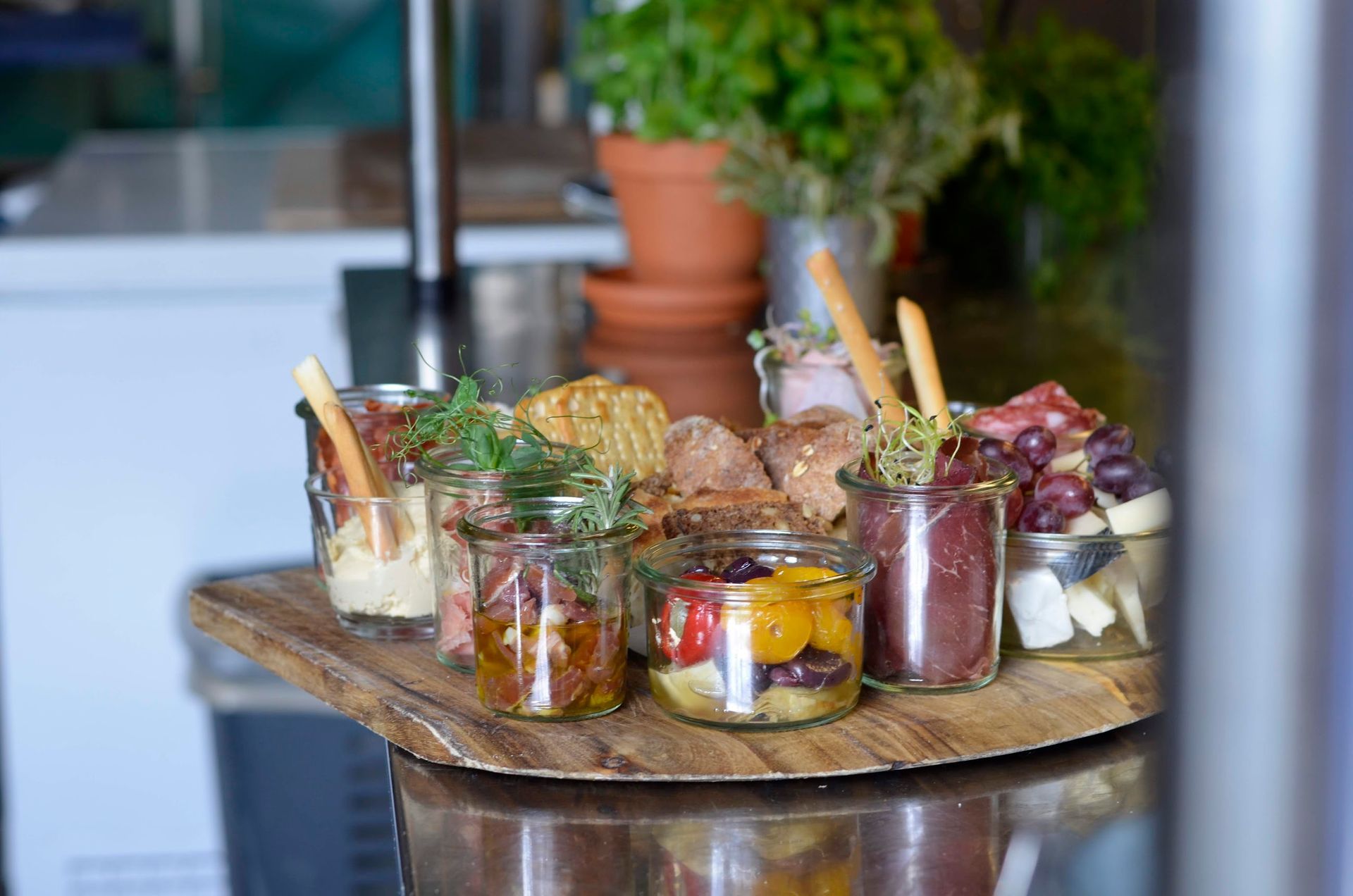 En række forretter serveret i små glaskrukker på et træfad med en potteplante i baggrunden.