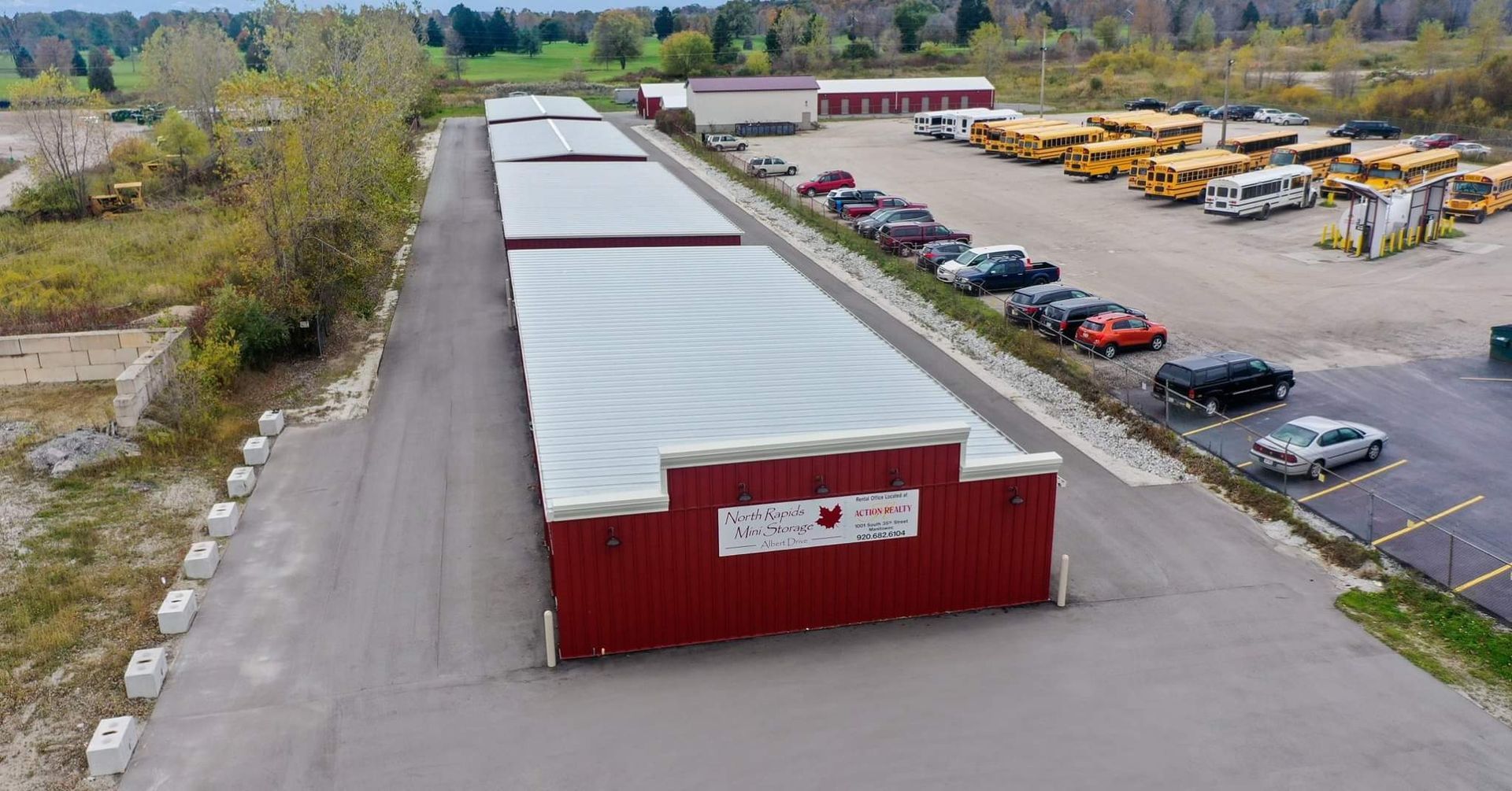 Red storage units, vehicles, and school buses on a paved lot. Overcast sky, rural setting.
