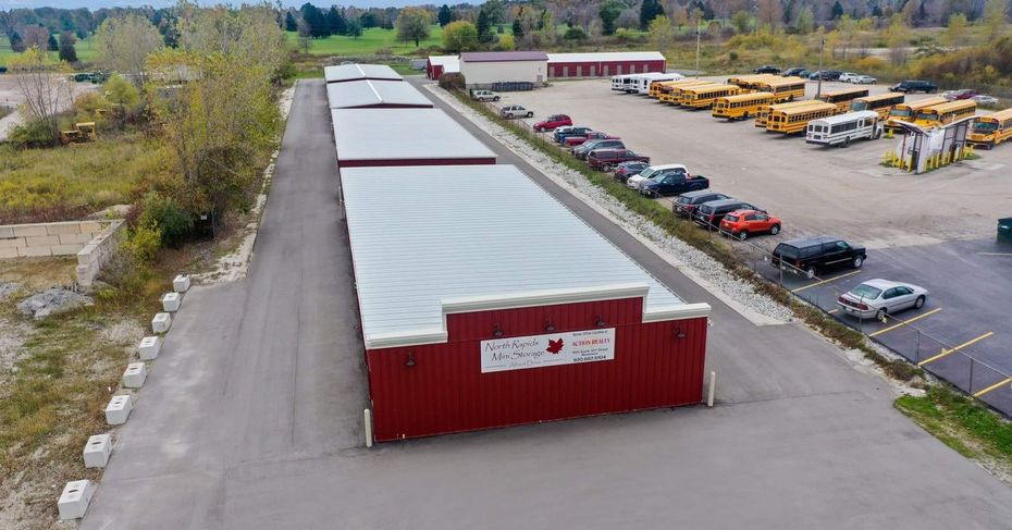 Red storage units, cars, and school buses in a paved lot. Buildings and trees in the background.