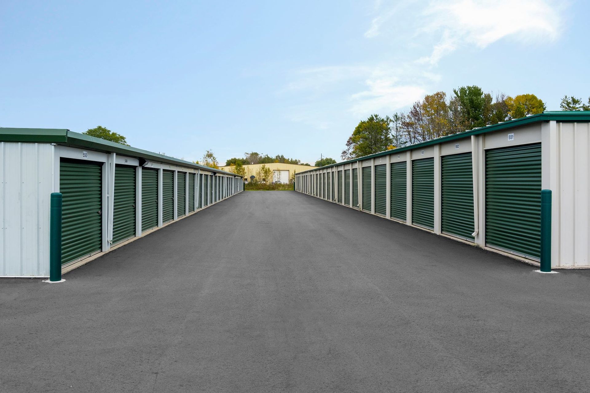 Storage units in a row. White doors with green accents line a gray asphalt drive under a blue sky.