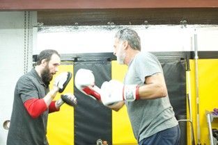 Two men boxing, one wearing gloves, the other holding pads. Gym setting.