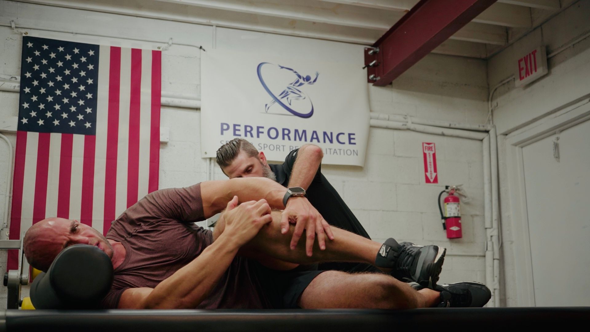 Man lying on a table, leg stretched as a trainer assists. American flag and a sign are in the background.