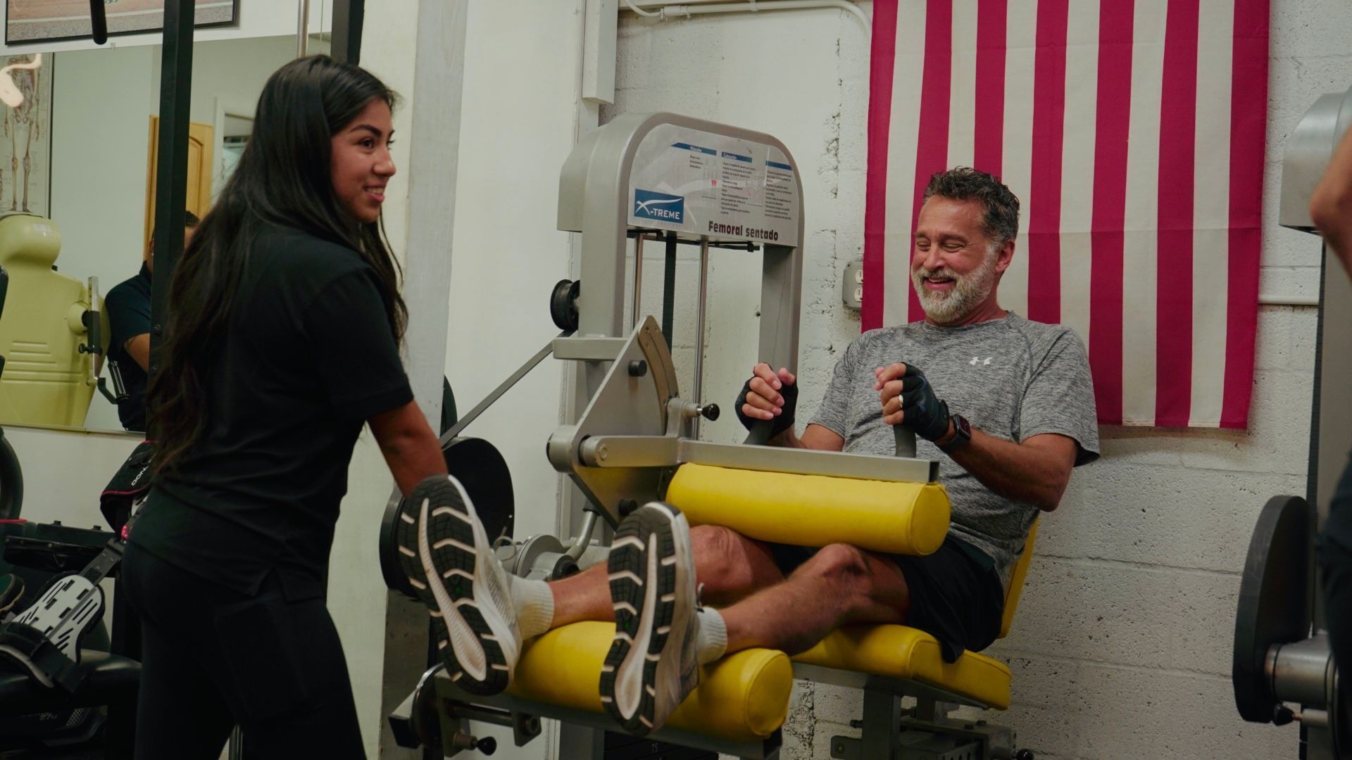 Woman assists man on yellow exercise machine in gym, with an American flag in the background