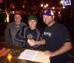 Three men at a table, two shaking hands over a document. One wears a Ravens hat. Dimly lit outdoor setting.