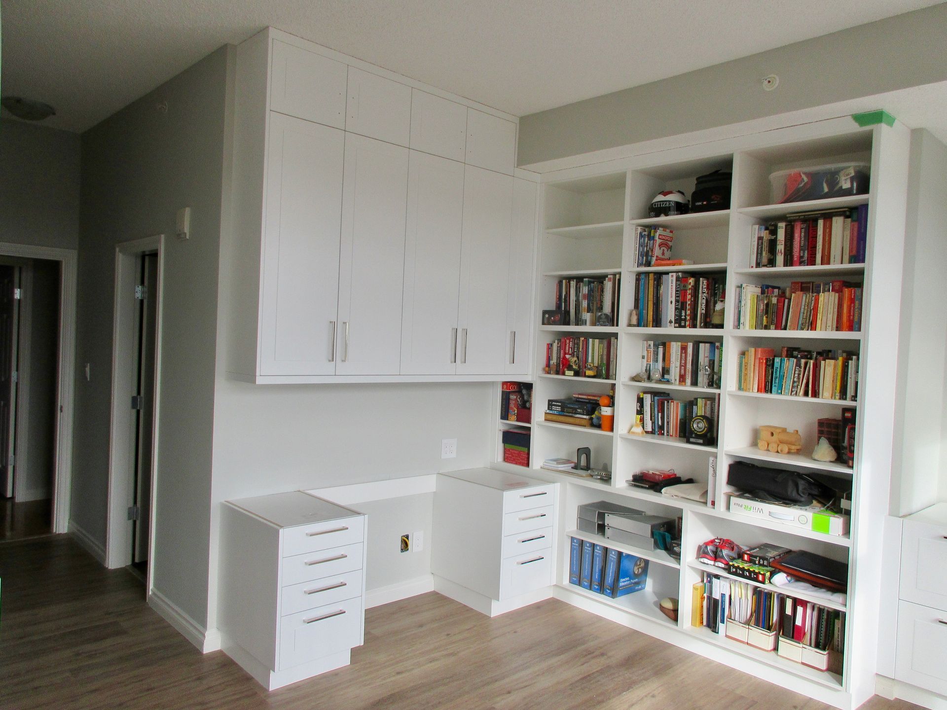 Built-in white shelves and desk unit in a corner. Cabinets above. Wooden floor and gray walls.