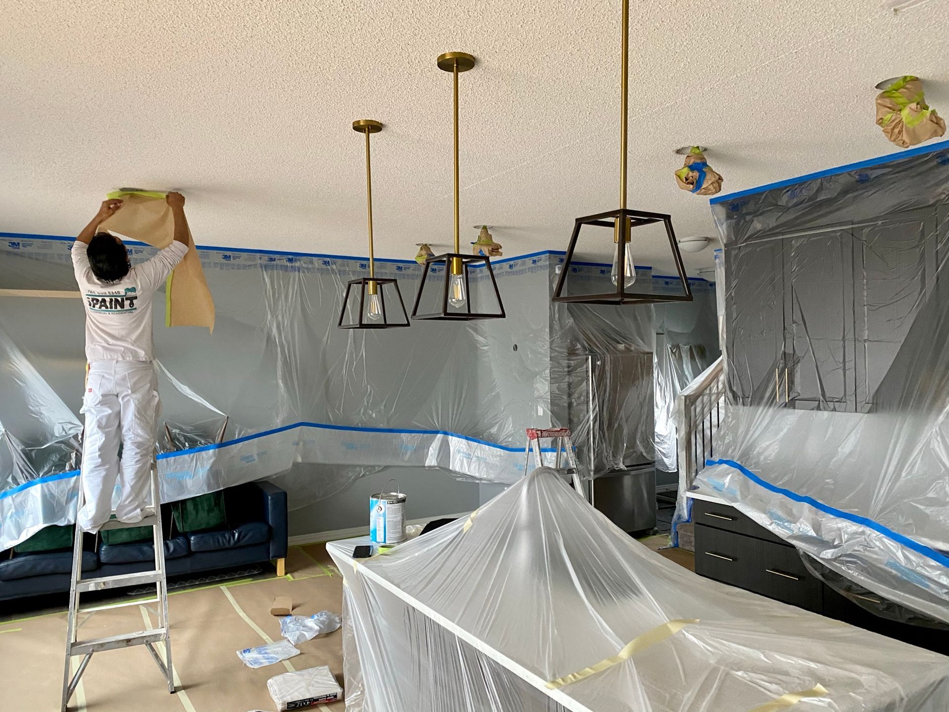 A man is standing on a ladder painting the ceiling of a living room.