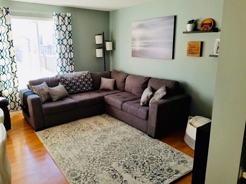 A living room with a brown sectional sofa and a patterned rug. Blue-green walls, window curtains, and decorative shelves.
