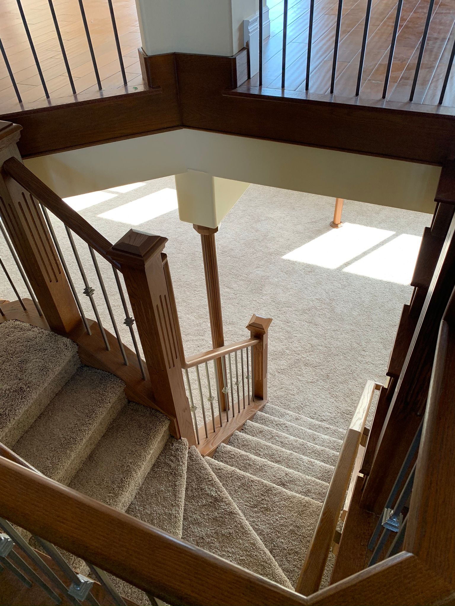 Staircase with wooden banisters and carpeted steps, viewed from above. Sunlight streams in from a window.