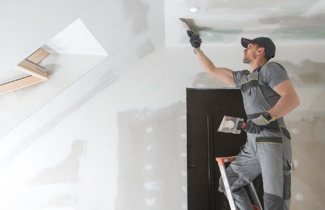 A man is standing on a ladder and plastering a ceiling.