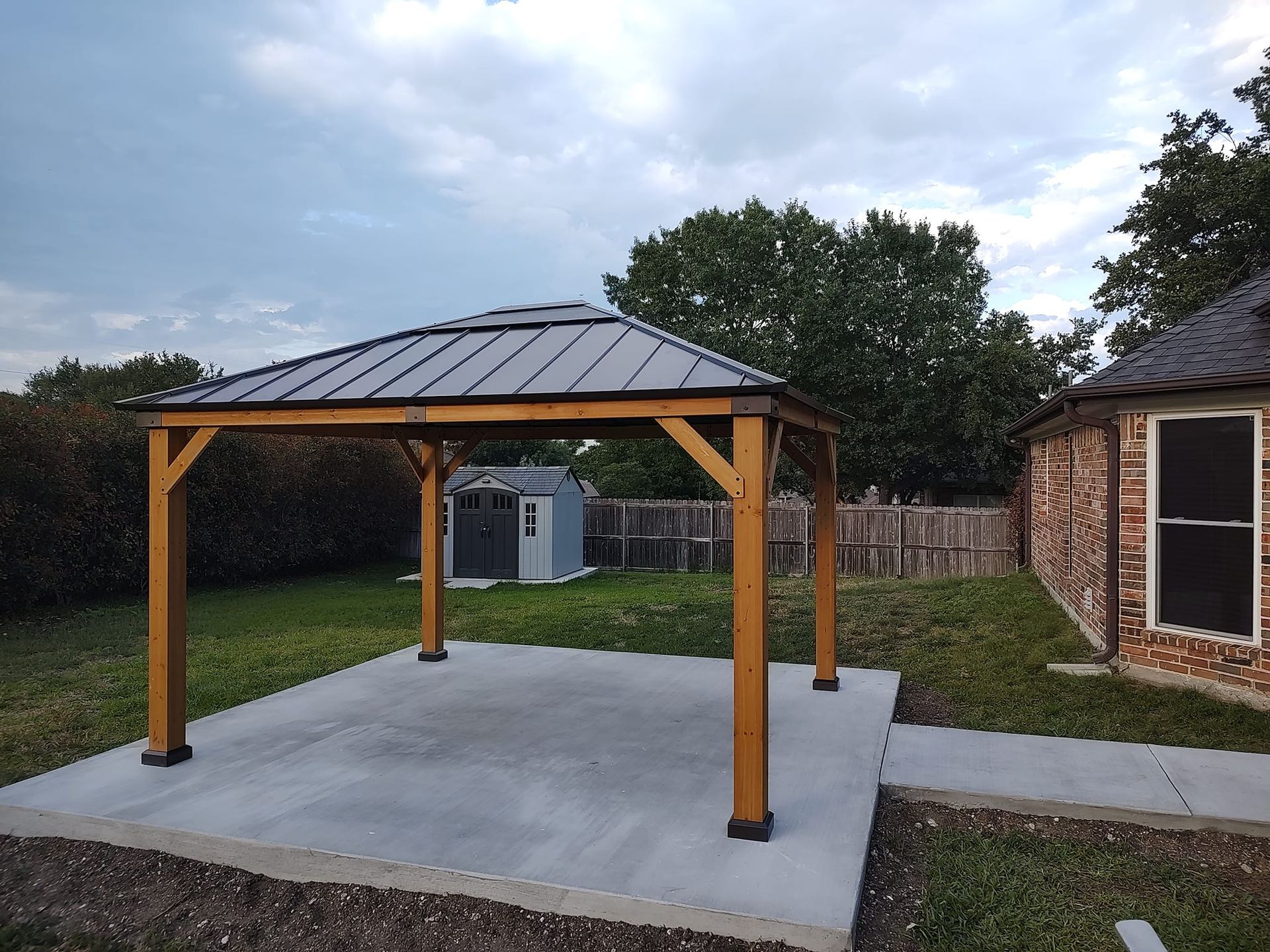 A wooden gazebo with a metal roof is in the backyard of a house.