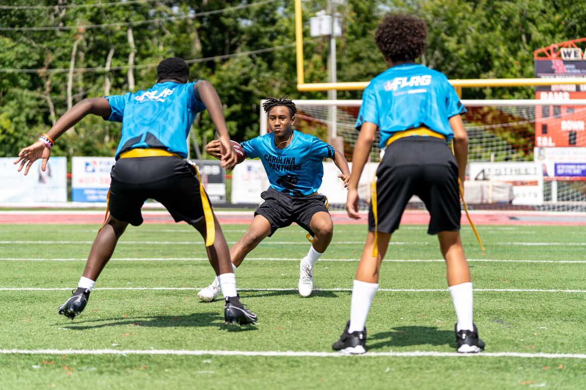 Football players in action on a grassy field; one runs with the ball, pursued by two opponents.