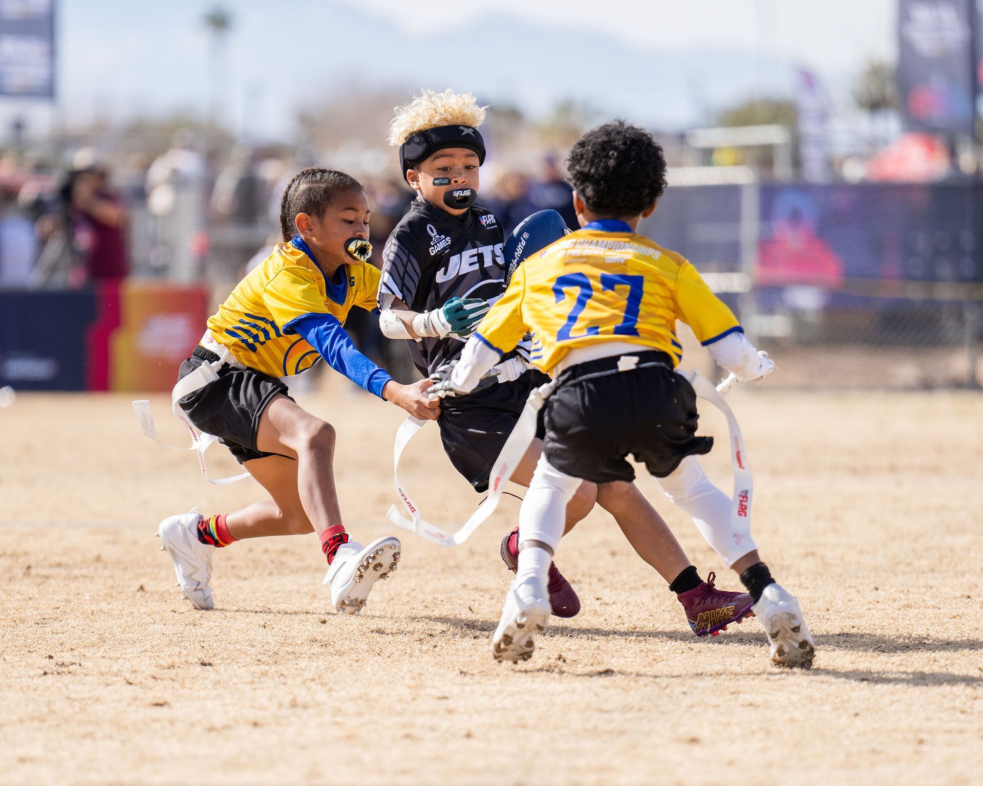 Football players in action on a grassy field; one runs with the ball, pursued by two opponents.